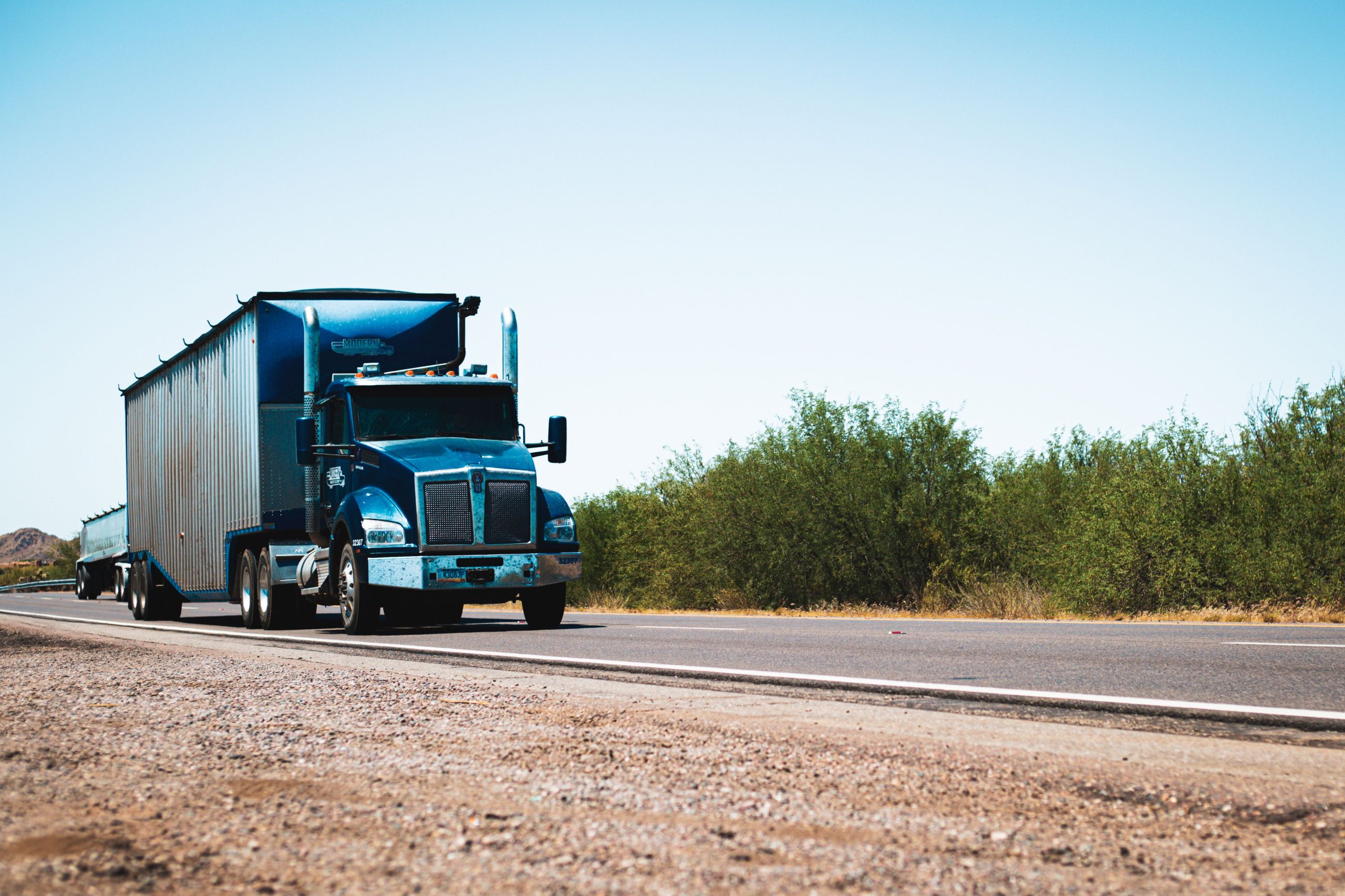 Blue semi-truck driving on a rural highway under clear blue skies